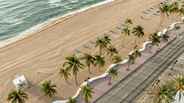 Ein Strand mit gepflastertem Gehweg, der von Palmen gesäumt ist, mit blauen Liegestühlen.
