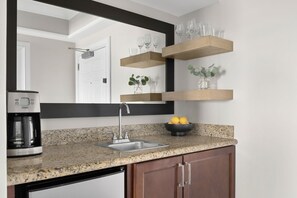 A kitchen with a granite countertop, stainless steel sink, and wooden cabinets. There are floating shelves with glassware and a bowl of lemons. - Dream Florida Escape in Fort Lauderdale! (Fort Lauderdale)
