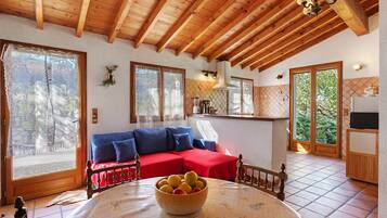 A cozy room with a wooden ceiling, a red sofa, a table with a fruit bowl, and a kitchen area with a refrigerator.