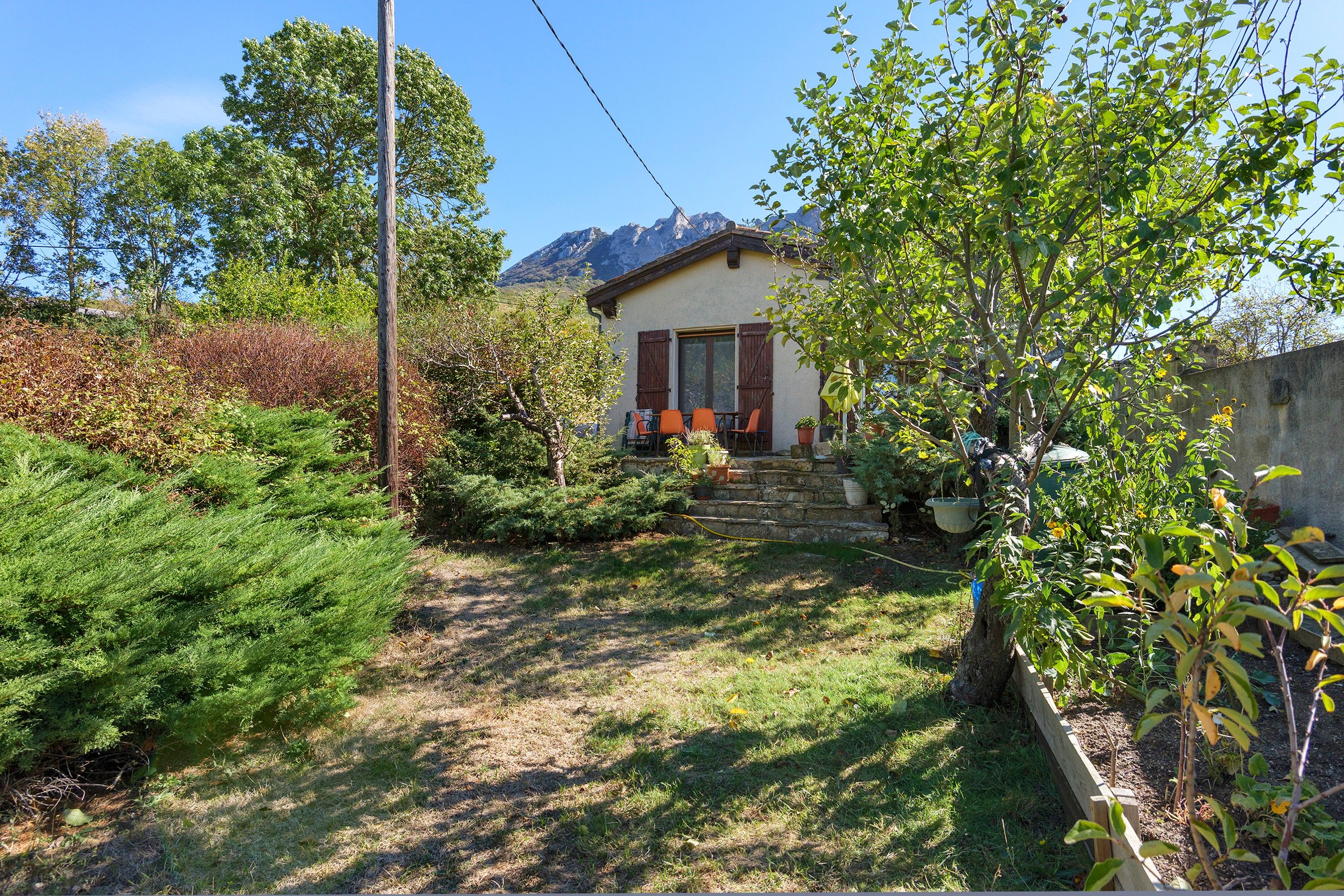 A house with a stone staircase leading to a wooden door, surrounded by greenery and a mountain in the background.