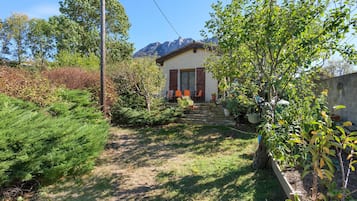 A house with a stone staircase leading to a wooden door, surrounded by greenery and a mountain in the background.