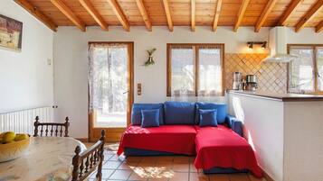 Une pièce chaleureuse avec un plafond en bois, un canapé rouge et bleu, une table à manger avec un saladier de fruits et un coin cuisine équipé d’une cuisinière.
