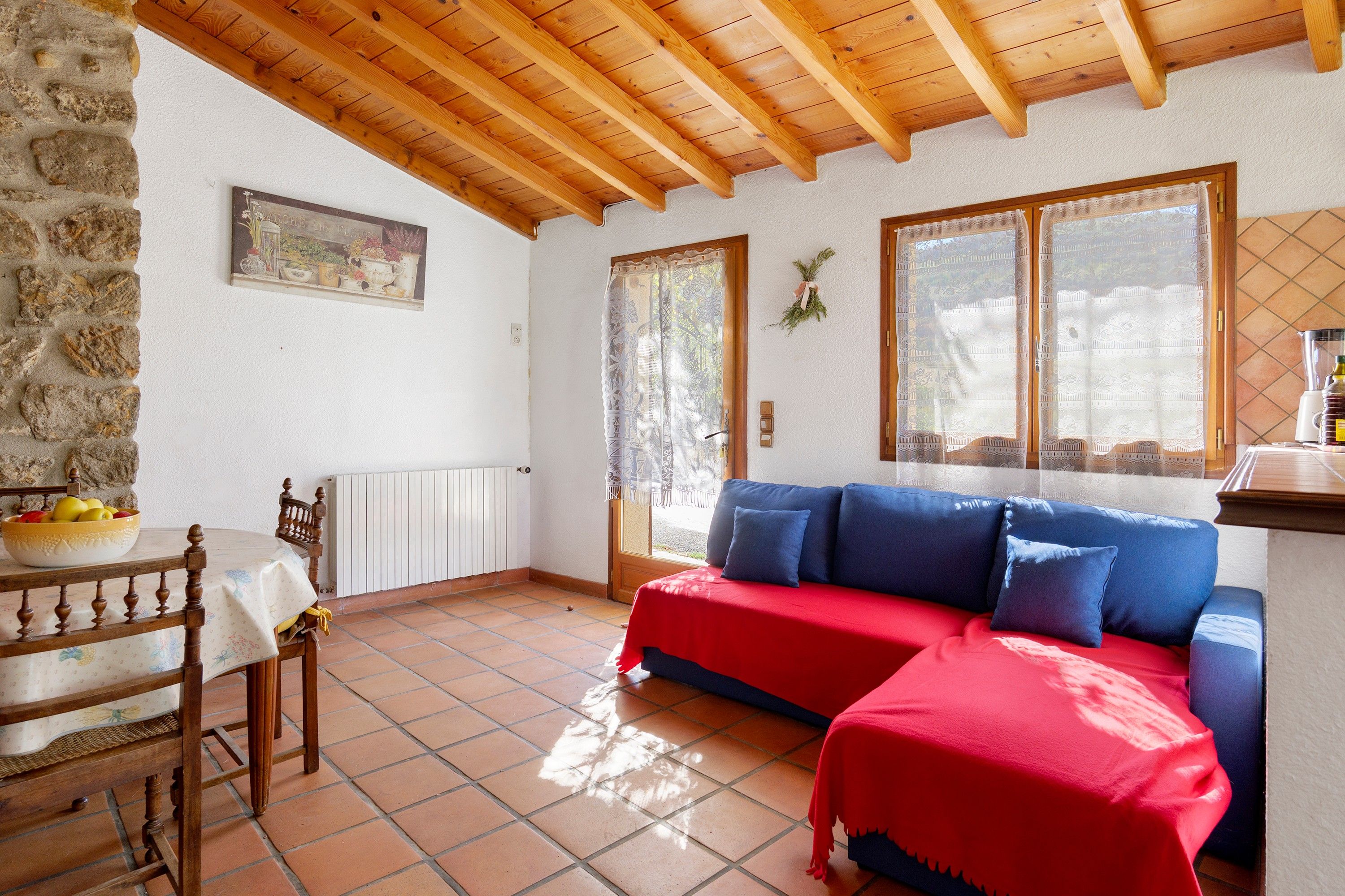 A cozy room with a wooden ceiling, a stone wall, a red sofa, and a dining table with a bowl of fruit.