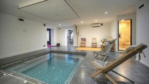 A modern indoor swimming pool with a clear water feature, surrounded by lounge chairs and a small seating area. - Exceptional 9-Bedroom Villa in Mougins (Mougins)
