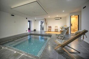 Ein moderner, überdachter Swimmingpool mit einer klaren Wasserattraktion, umgeben von Liegestühlen und einer kleinen Sitzgelegenheit.
