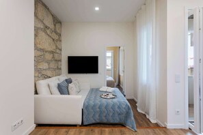 Ein modernes Schlafzimmer mit Steinwand, Flachbildfernseher, weißem Sofa, gemustertem Bettbezug und Holzboden.