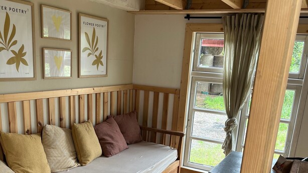 A cozy room with a wooden bench, a cushioned bench, and a window with curtains. - Big Break in the Tiny House on the Farm Near the Baltic Sea (Schashagen)