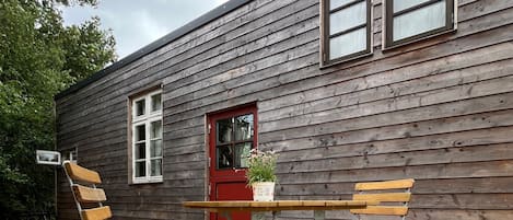 A wooden deck with a table and two chairs, a red door, and a window.