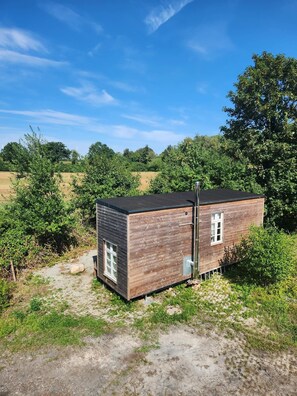 A small wooden cabin with a metal chimney, surrounded by greenery and trees.