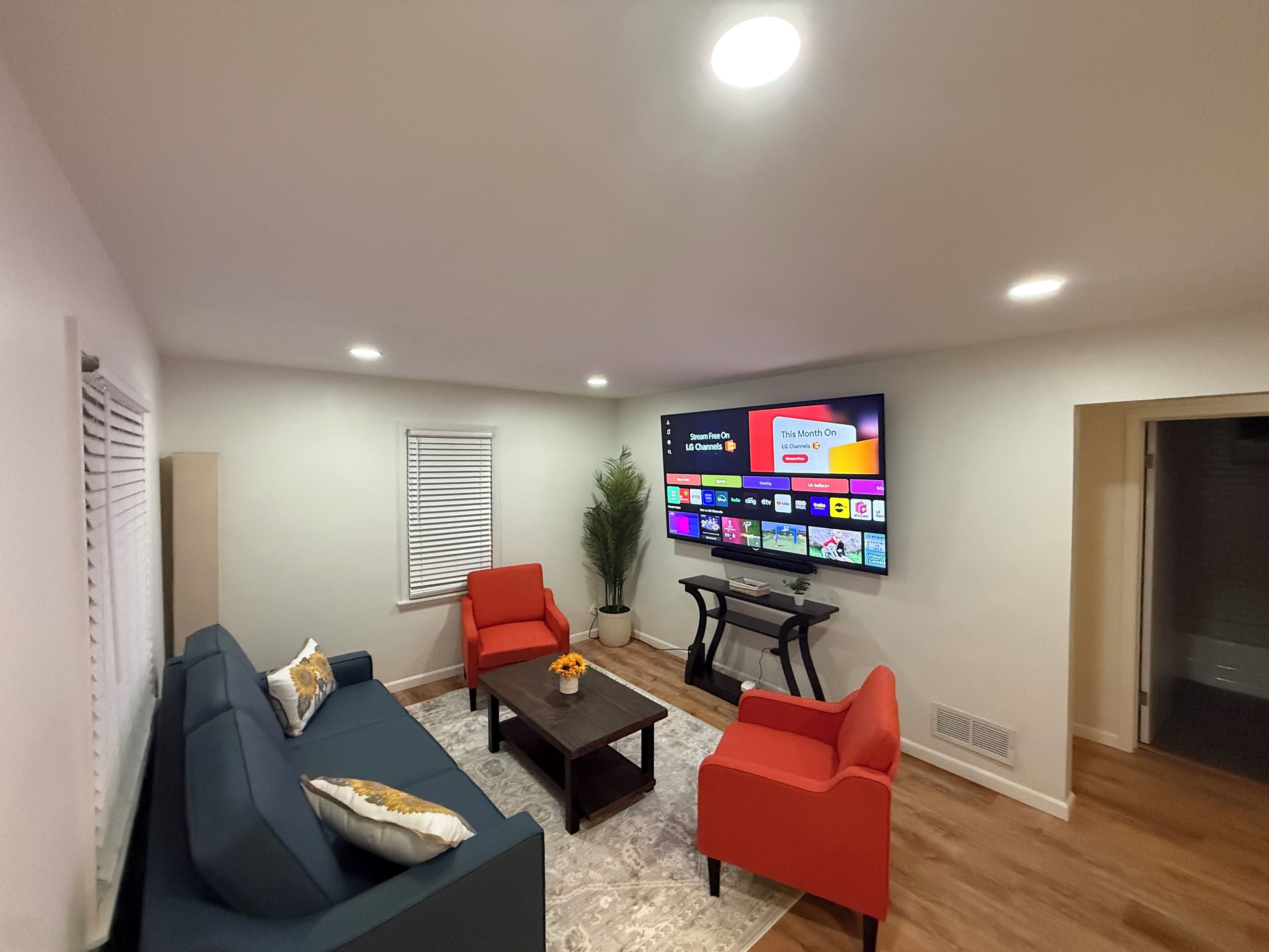 Una sala de estar moderna con una televisión de pantalla plana en la pared que muestra varios canales, un sofá azul, un sillón rojo y una mesa de centro con un jarrón.