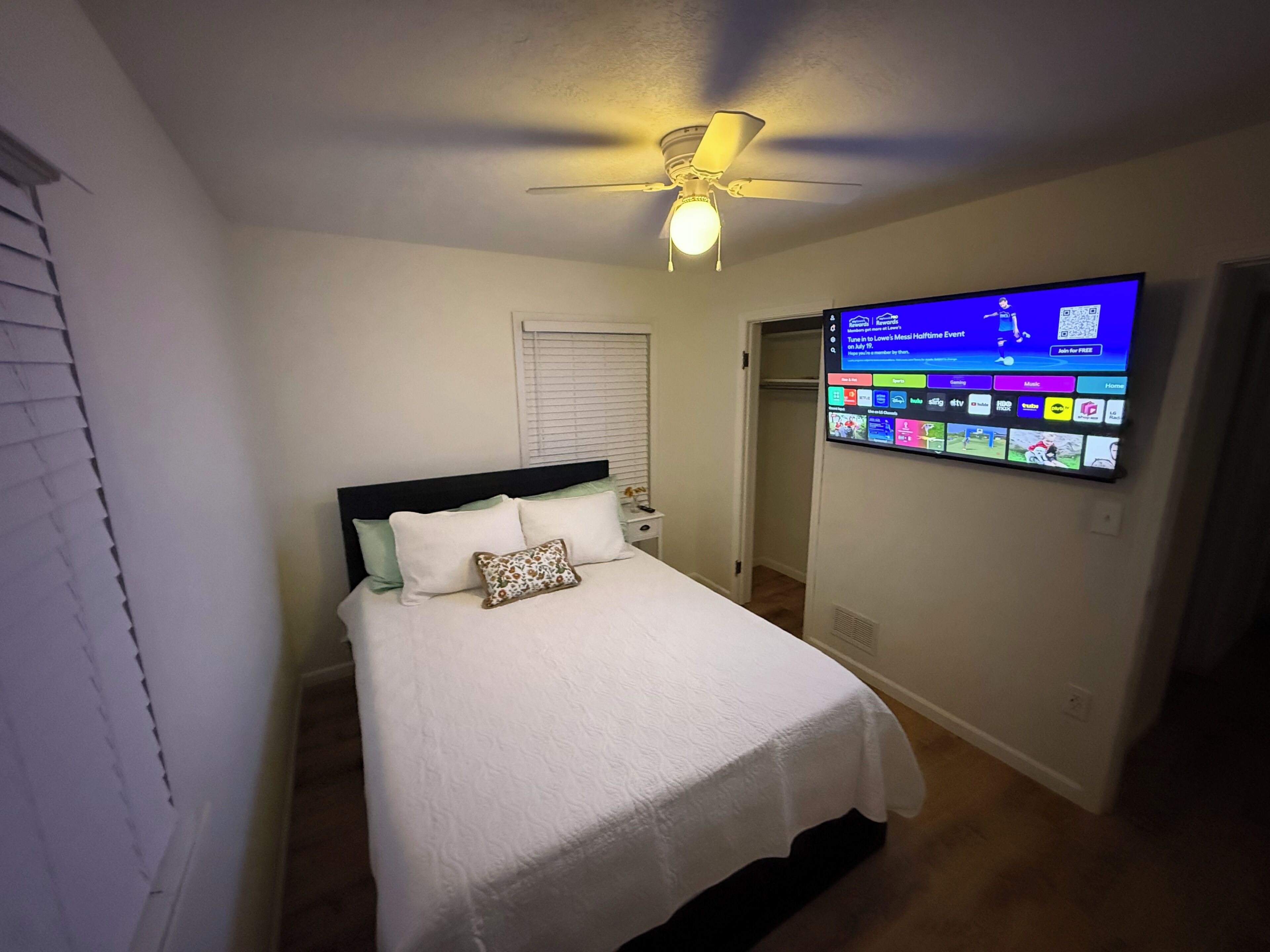 Un dormitorio con una cama, ventilador de techo y televisor en la pared mostrando un canal deportivo.
