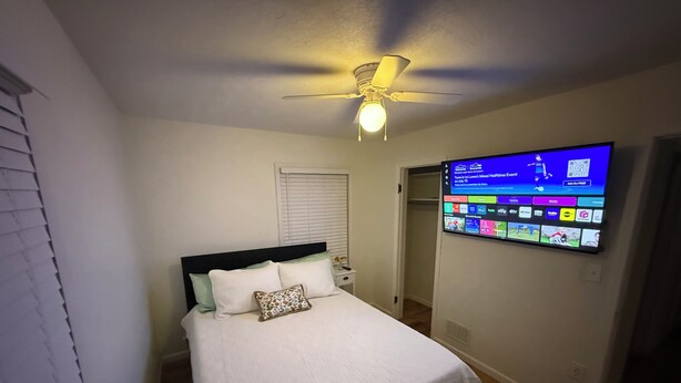 Une chambre à coucher avec un lit, un ventilateur de plafond et un téléviseur fixé au mur, diffusant une chaîne de sport.