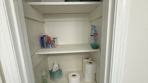 Un petit espace de rangement doté d’étagères où sont rangés des serviettes pliées, des produits de nettoyage et un panier contenant un objet rouge.