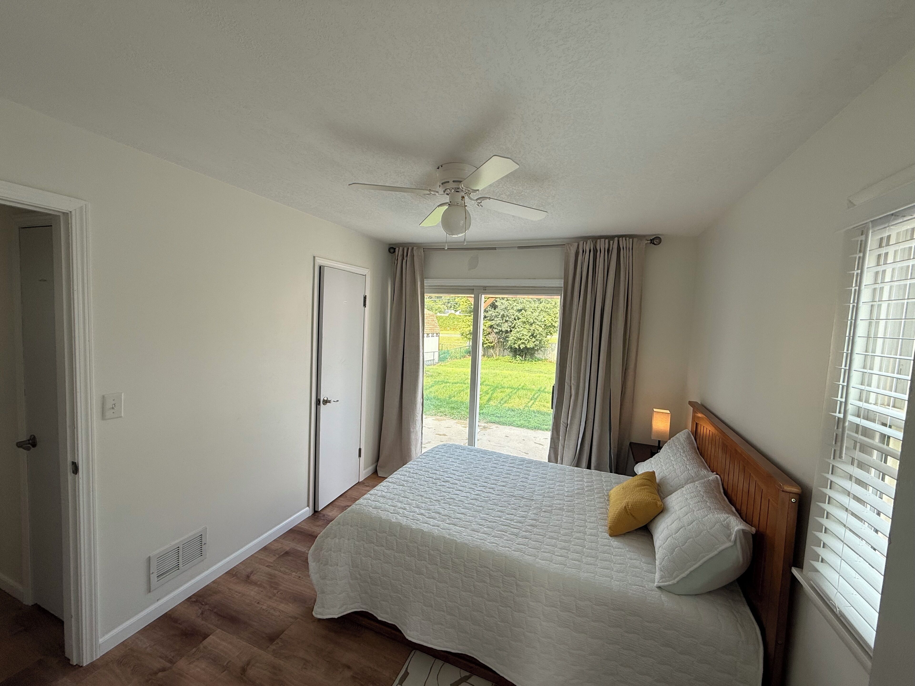 Un dormitorio con una cama, ventilador de techo, ventana con persianas y puerta que da a otra habitación.