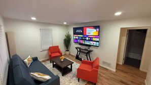 Un salon avec un téléviseur à écran plat fixé au mur, un canapé de couleur foncée, deux fauteuils rouges, une table basse et une plante en pot.