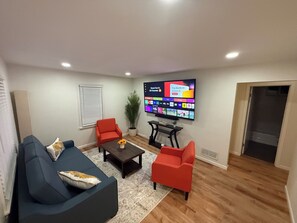 A living room with a flat-screen TV mounted on the wall, a dark-colored sofa, two red chairs, a coffee table, and a potted plant. - VIP Suite—2BR/1 BA House Near Mayo Clinic Hospital (Rochester)