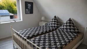 Ein Zimmer mit einem Einzelbett, einer geometrisch gemusterten Bettdecke, einem weißen Nachttisch mit Lampe und einem gerahmten Bild an der Wand.