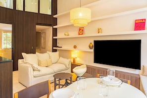 A modern living room with a sofa, a dining table with glasses and cutlery, a TV mounted on the wall, and a bookshelf with decorative items. - Modern 2bdr Flat for 6P - Saint-tropez / Port (Saint-Tropez)