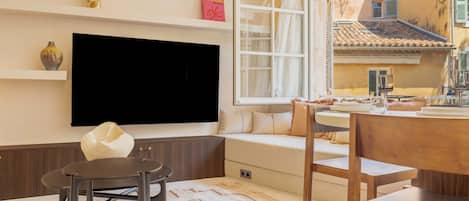 A modern living room with a flat-screen TV, a sofa, a dining table, and a window overlooking a building.