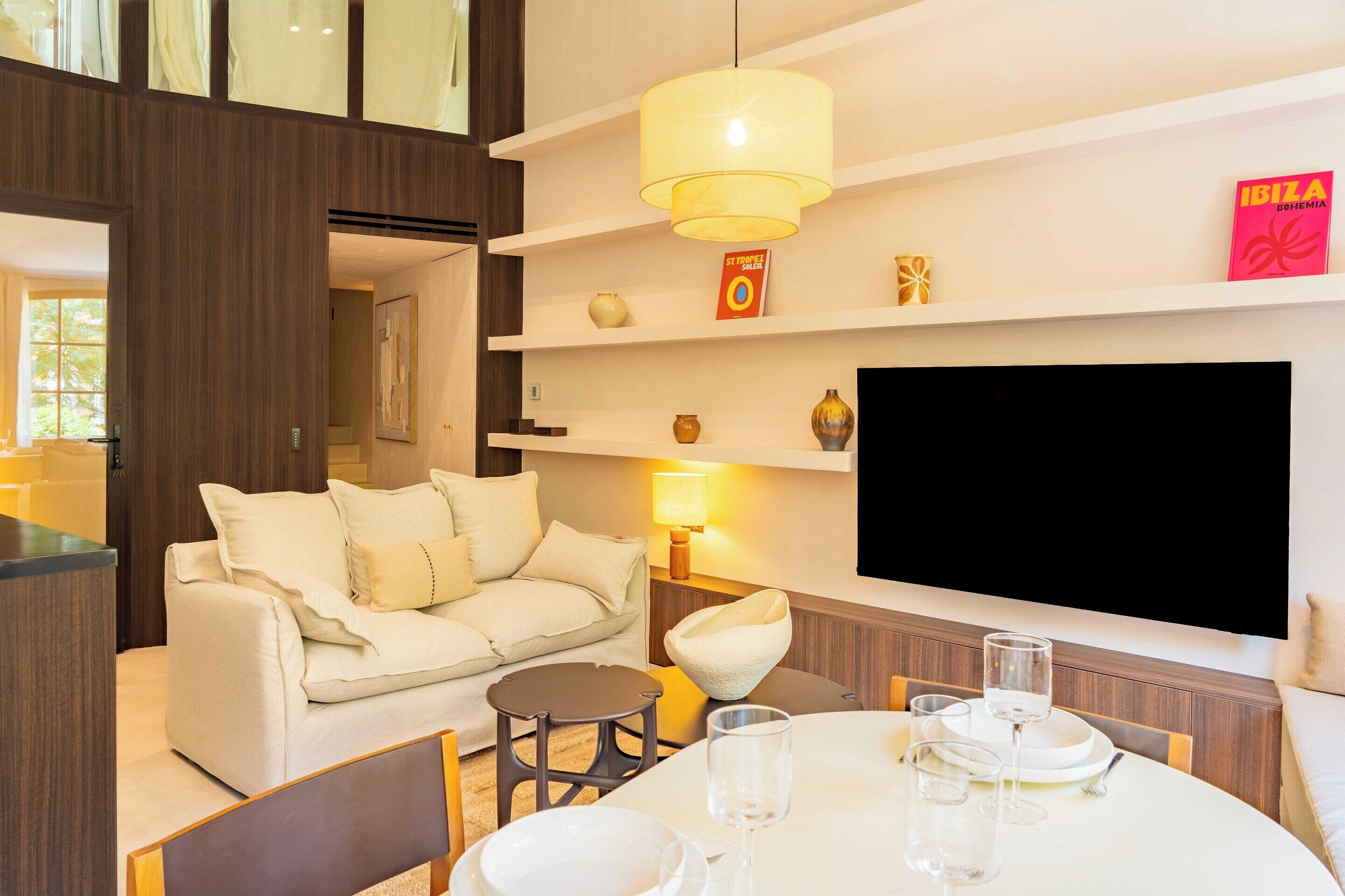 A modern living room with a sofa, a dining table with glasses, a TV mounted on the wall, and a bookshelf with decorative items.