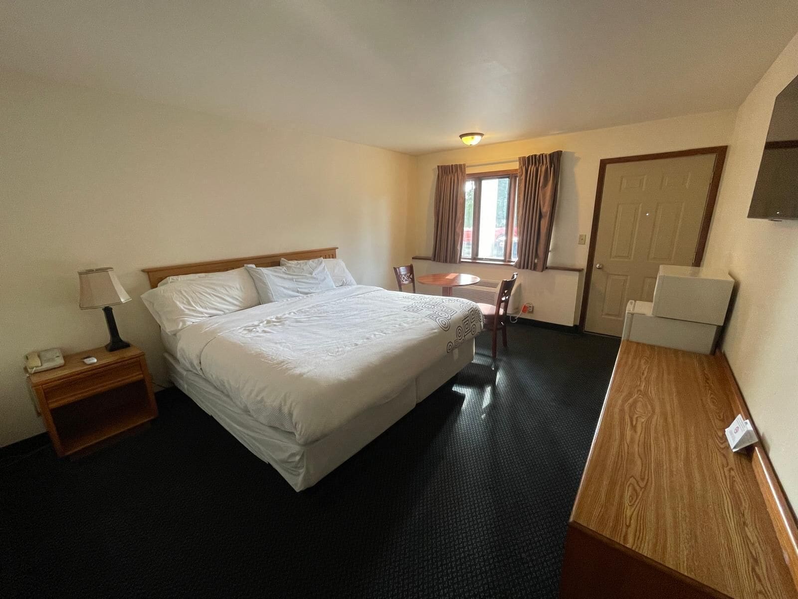 Quarto de hotel com cama, mesa de cabeceira, luminária, cadeira, escrivaninha e porta.