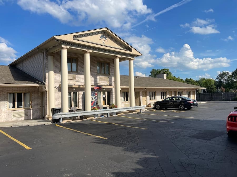 A building with a central entrance flanked by columns, a parking lot, and a red vehicle in the foreground.
