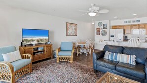 A living room with a television, a blue sofa, a wicker chair, a dining table with chairs, and a kitchen area with cabinets and appliances. - Oceanside 205-Private Balcony Sunsets (Indian Rocks Beach)