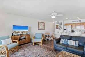Ein Wohnzimmer mit Fernseher, einem blauen Sofa, einem Korbstuhl, einem Esstisch mit Stühlen sowie einer Küchenzeile mit Schränken und Geräten.
