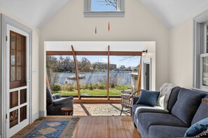 Ein Zimmer mit großem Fenster mit Blick auf ein Gewässer, einer Holzschaukel, einer Couch und einem gemusterten Teppich.