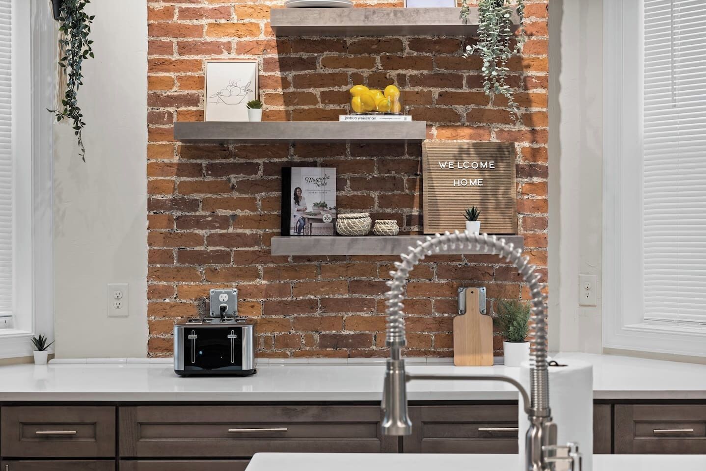 A kitchen with a brick wall, a white countertop, a built-in sink, and a shelf with decorative items.