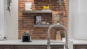 A kitchen with a brick wall, a white countertop, a built-in sink, and a shelf with decorative items.
