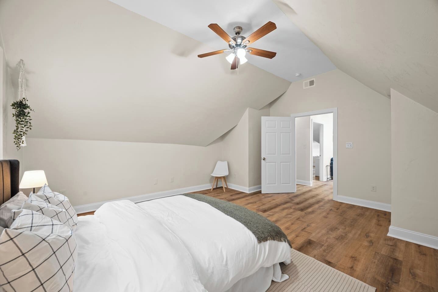 Une chambre avec un plafond en pente, un ventilateur de plafond, un lit avec une literie blanche, une table de chevet avec une lampe et une porte donnant sur une autre pièce.