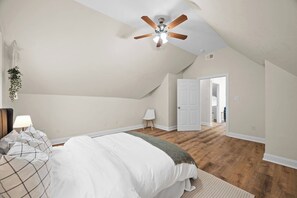 Ein Schlafzimmer mit schrägem Dach, einem Deckenventilator, einem Bett mit weißer Bettwäsche, einem Nachttisch mit Lampe und einer Tür zu einem weiteren Raum.