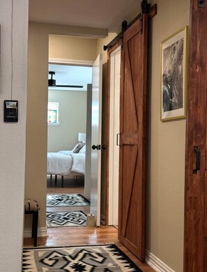 A hallway with a wooden door, a bed in the adjacent room, and a patterned rug on the floor. - Walk to Dickies&Will Rogers|Guest Suite+Game Room (Fort Worth)