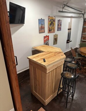 A bar area with a wooden counter and stools, a television mounted on the wall, and vintage posters on the wall. - Walk to Dickies&Will Rogers|Guest Suite+Game Room (Fort Worth)