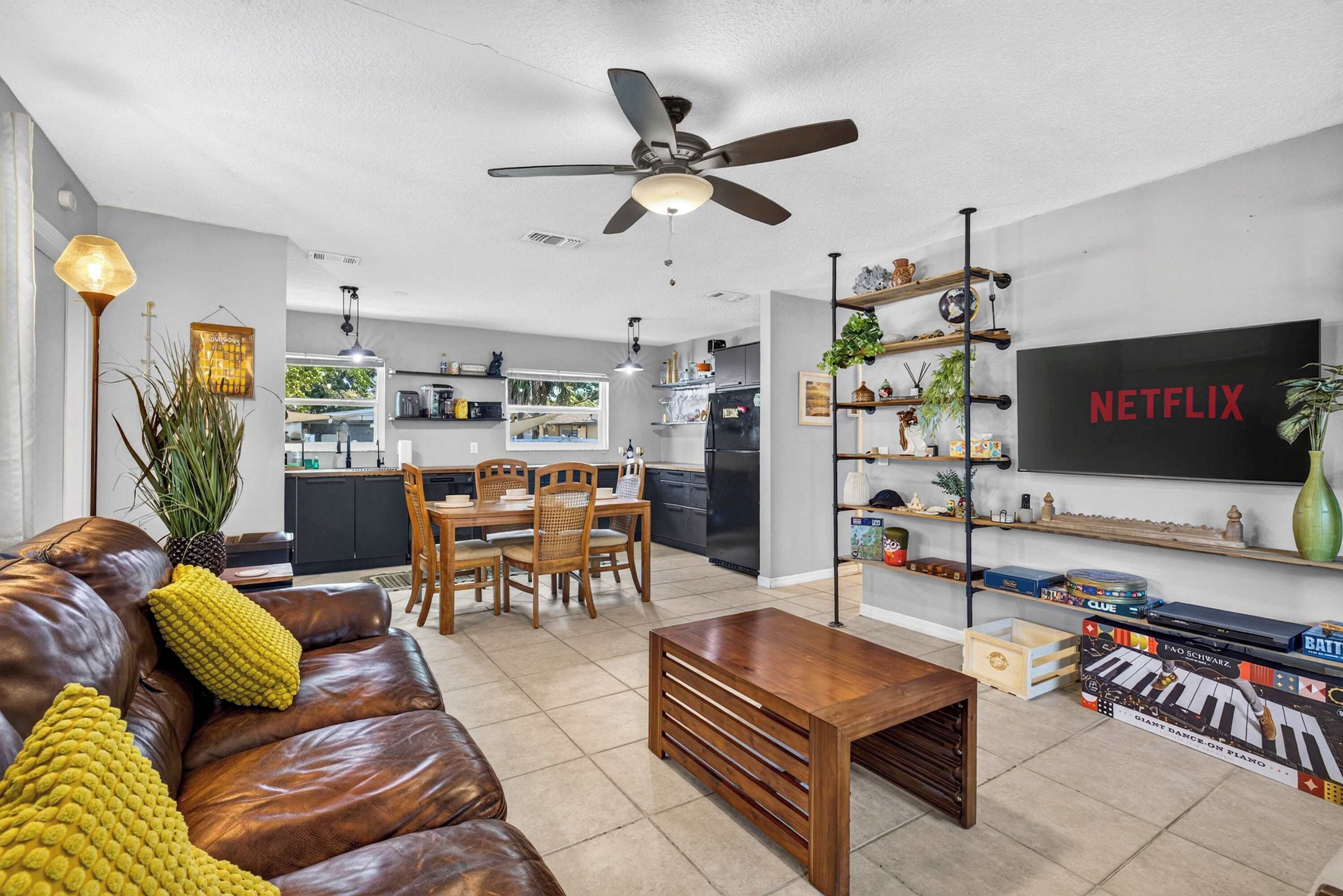 Een moderne woonkamer met een leren bank, een houten salontafel, een eethoek met een houten tafel en stoelen, een keuken op de achtergrond, en een tv met daarop 'NETFLIX'.
