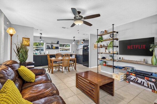 Un salon moderne avec un canapé en cuir, une table basse en bois, un coin repas avec une table et des chaises en bois, une cuisine en arrière-plan et un téléviseur fixé au mur qui affiche « NETFLIX ».