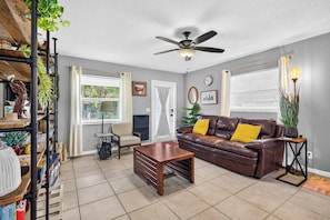 A living room with a brown leather sofa, a wooden coffee table, a ceiling fan, and a window with blinds. - The Landing Pad | Pet Friendly Yard (Tampa)