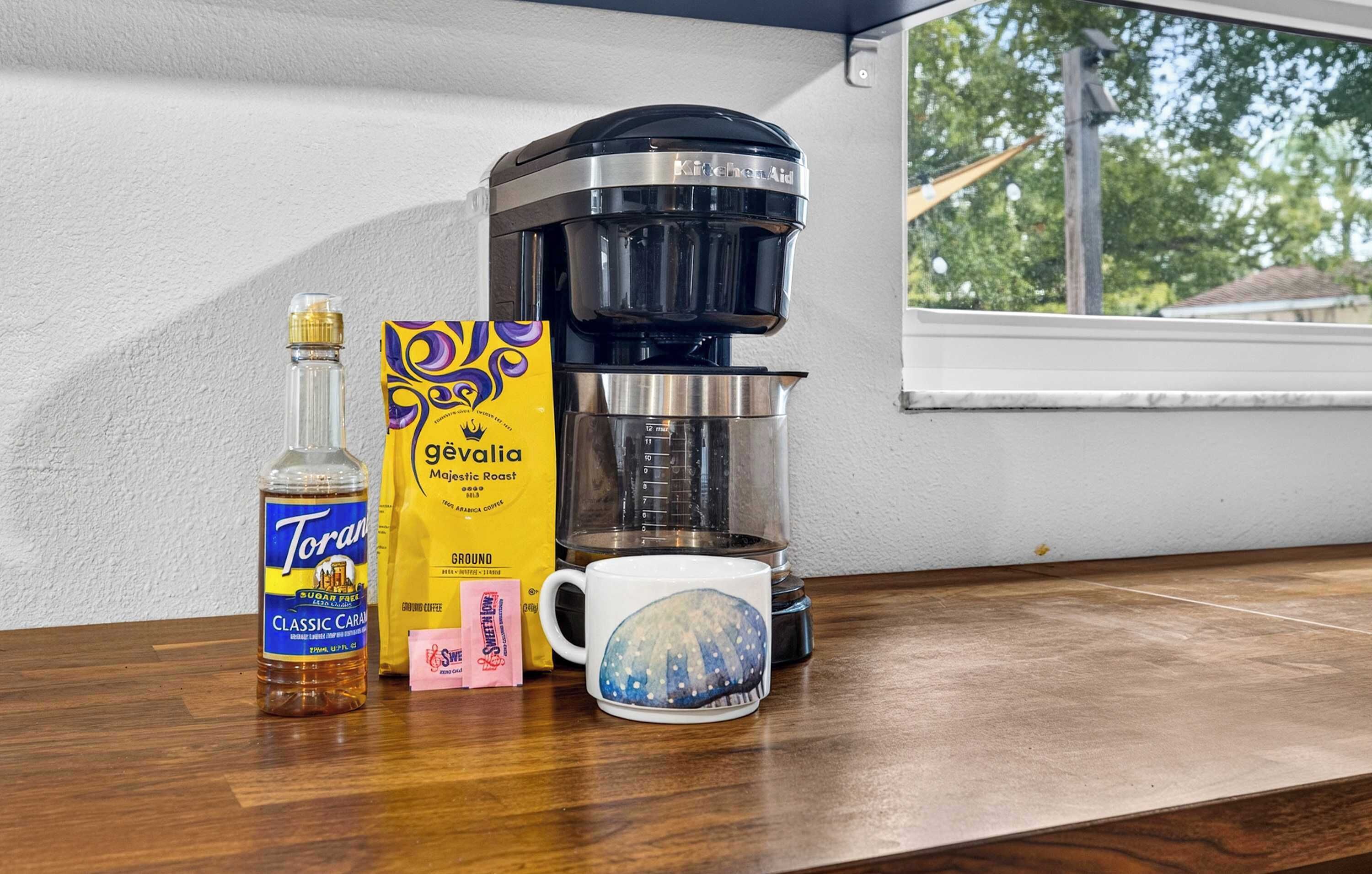 Une machine à café, une bouteille de sirop caramel classique de la marque Torani, un paquet de café Geyalga Majestic Roast, un paquet rose de café Swell, et une tasse ornée d’un motif bleu et blanc, le tout posé sur une table en bois.