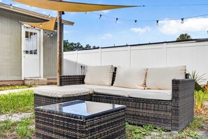 A wicker sofa with cushions, a glass-top coffee table, and a string of lights against a white wall. - The Landing Pad | Pet Friendly Yard (Tampa)