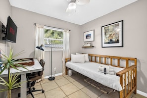 A small, well-lit room with a single bed, a desk with a chair, a standing lamp, a ceiling fan, and a wall-mounted TV. - The Landing Pad | Pet Friendly Yard (Tampa)