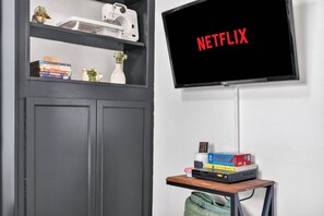 Une pièce avec un téléviseur affichant le mot « NETFLIX » en lettres rouges, une étagère avec des livres et une petite plante, et une table avec une boîte de jeux de puzzle et une télécommande.