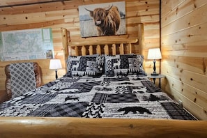Una habitación de cabaña de madera con una cama, dos mesitas de noche, un mapa en la pared y un cuadro grande de animales sobre la cama.