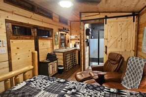 A cozy cabin interior with a bed, a seating area with leather chairs, a small kitchenette, and a sliding door leading to a shower area.