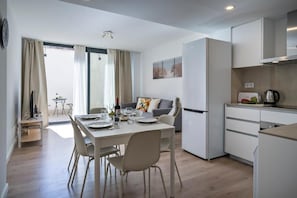 Een moderne keuken met witte kastjes, een eethoek met een tafel gedekt voor twee personen, en een groot raam met gordijnen.