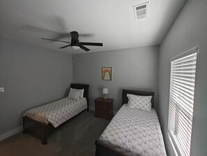 A bedroom with two single beds, a ceiling fan, a nightstand, a lamp, and a window with blinds.