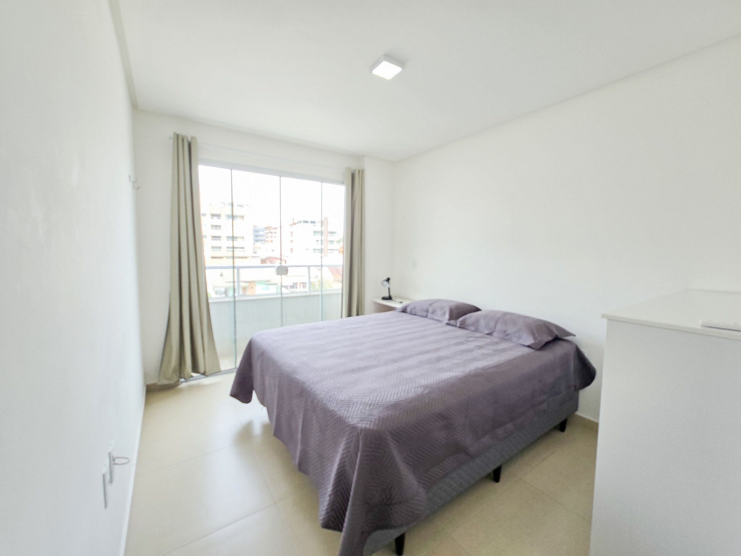 A bedroom with a bed, a nightstand, a window with curtains, and a view of buildings outside.