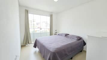 A bedroom with a bed, a nightstand, a window with curtains, and a view of buildings outside.