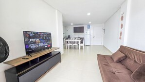 A living room with a flat-screen TV, a brown sofa, and a dining area with white chairs and a table. - La Rosadita by LikeHome  (Bombinhas)