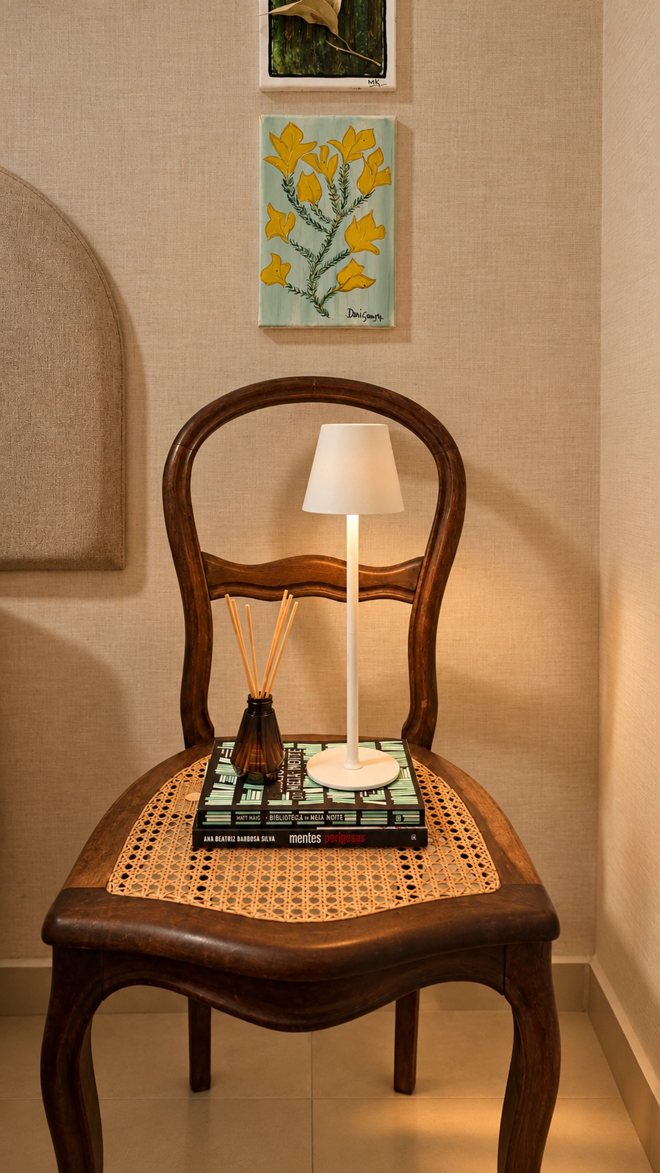 A wooden chair with a woven seat cushion, a white table lamp, and a book titled 'mentes perigosas' on a table.